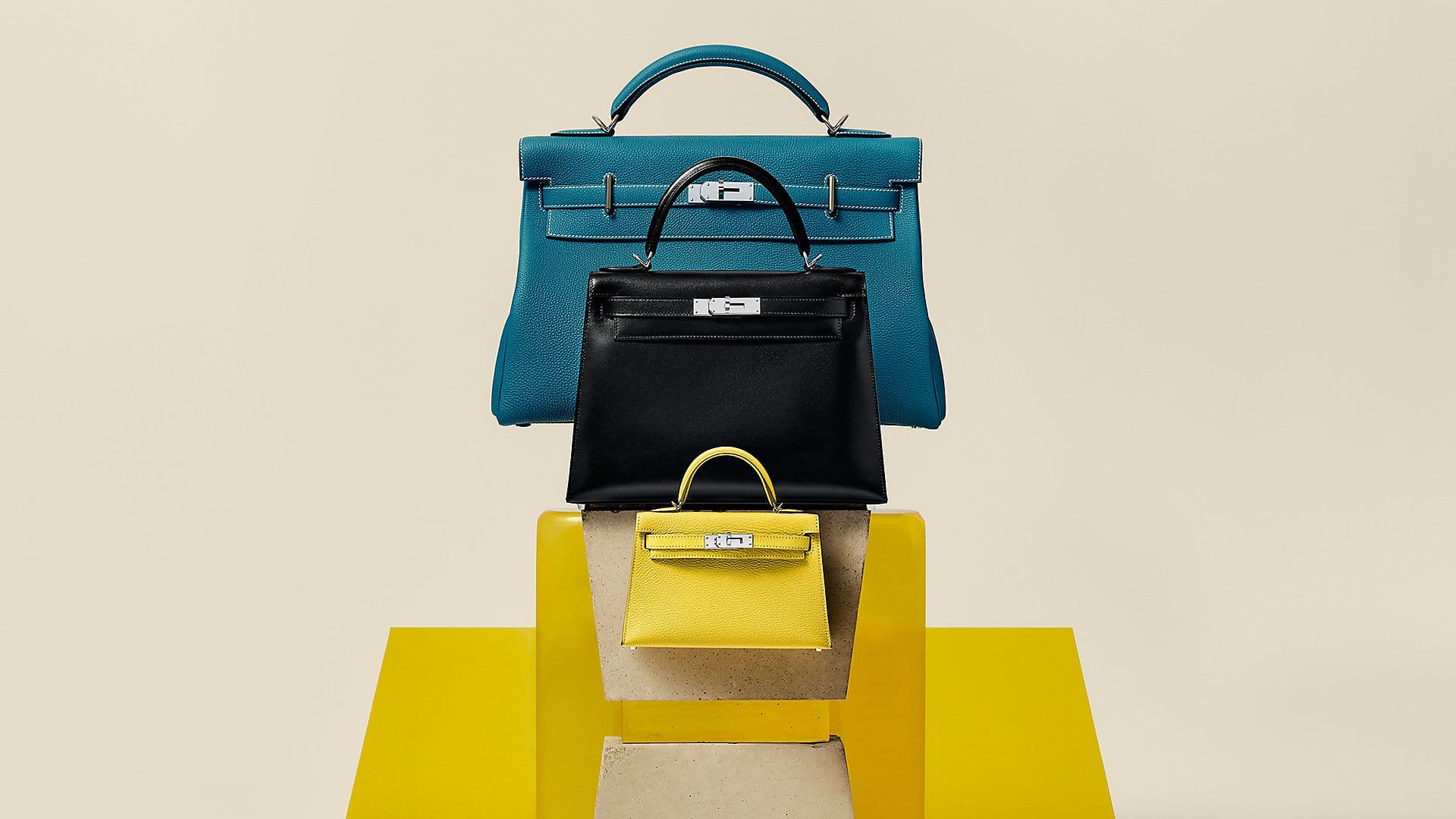 Three Hermès Kelly bags in blue, black and yellow arranged vertically on geometric plinths.