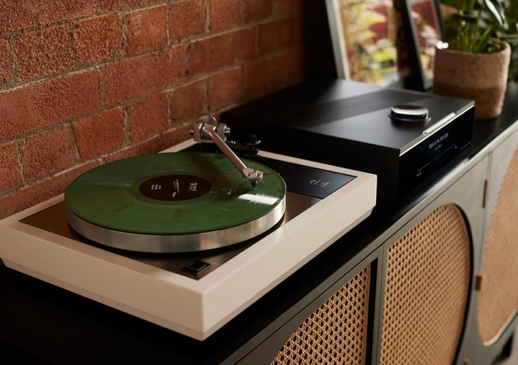 A white-plinth Linn Selekt LP12 playing a green record on a dark cabinet beside a brick wall.