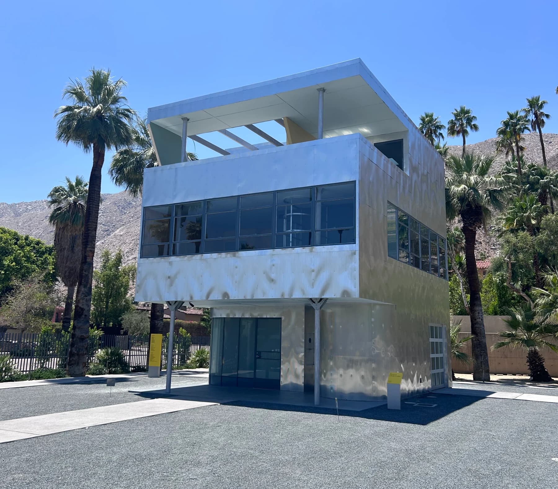 The reconstructed silver Aluminaire House in Palm Springs, with its raised rectangular volume, slender supports and palm trees against a clear blue sky.