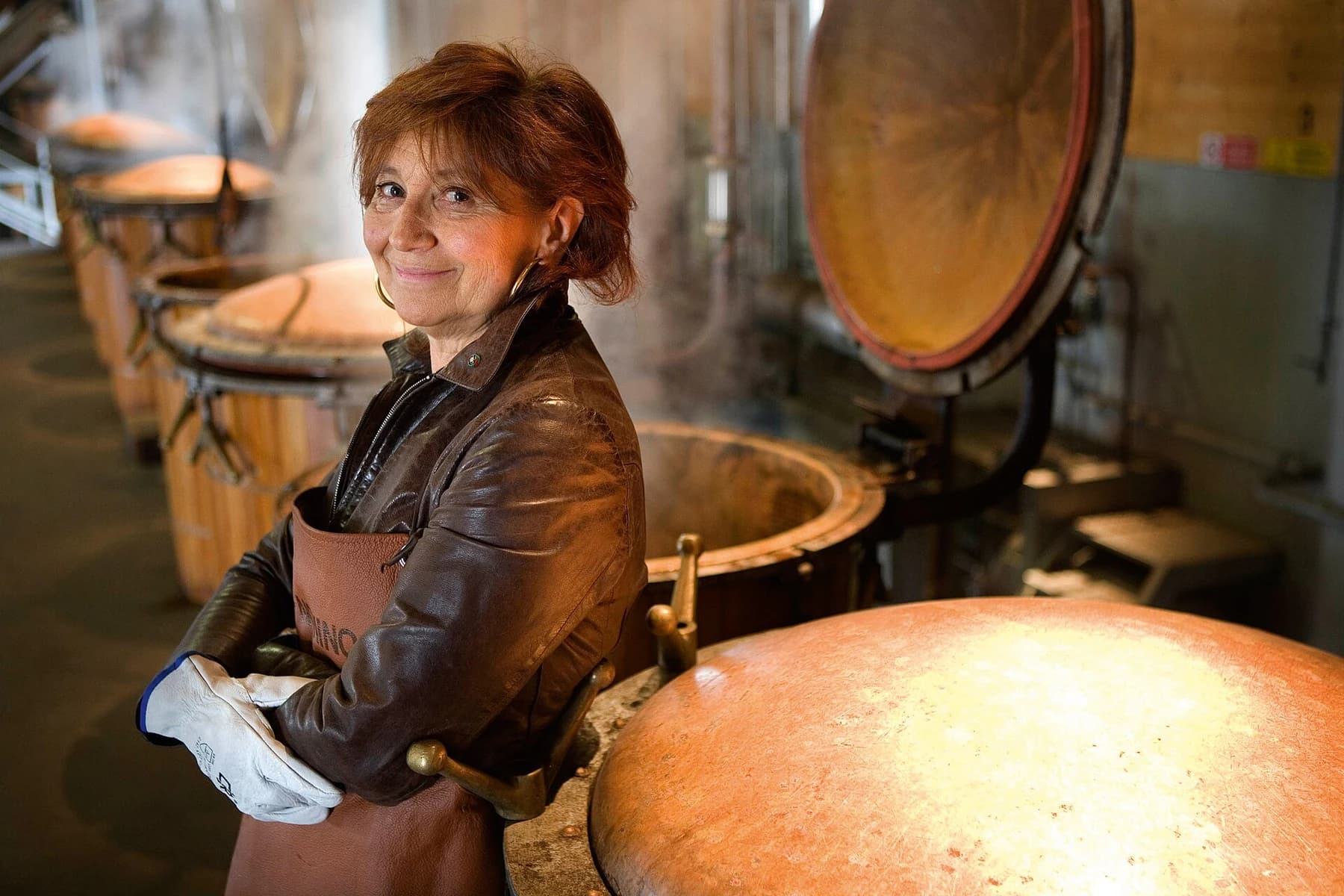 Giannola Nonino smiling beside polished copper distillation equipment, wearing a brown jacket and holding work gloves.