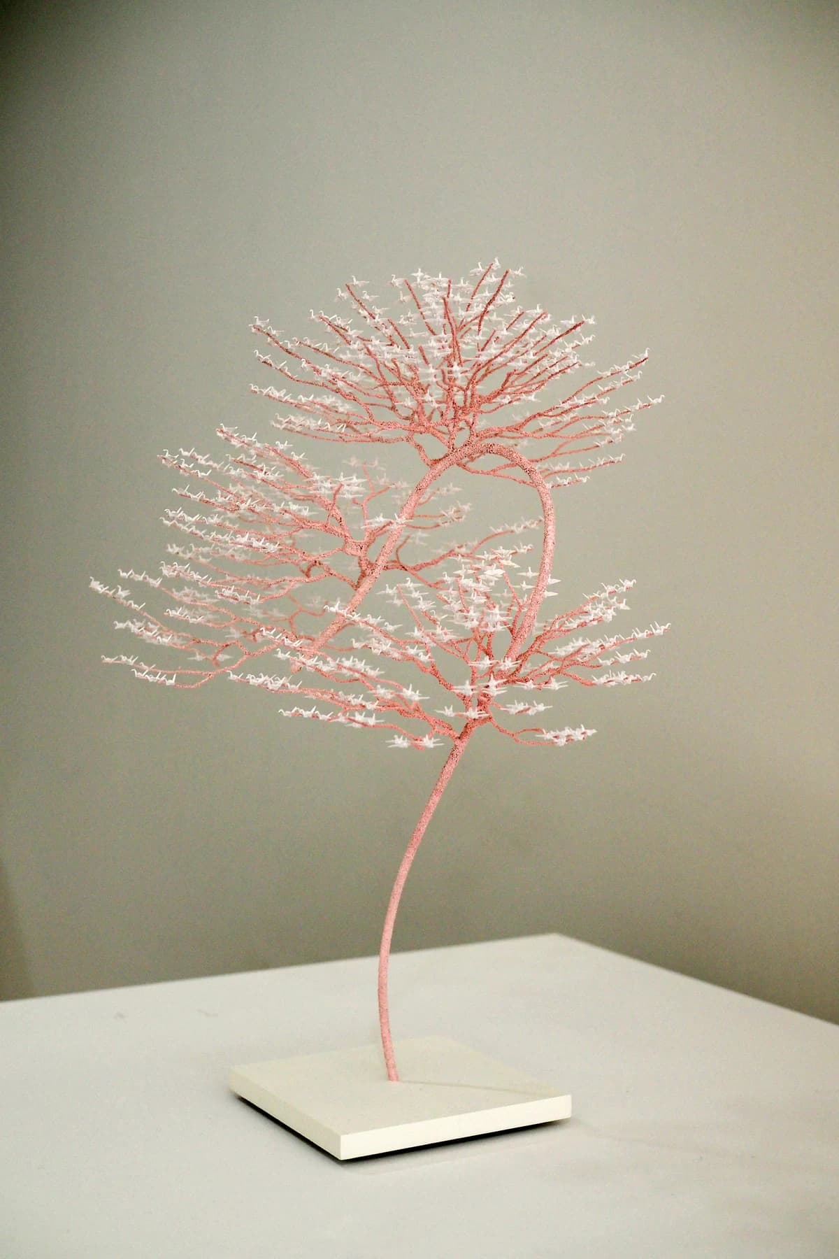 A delicate tree-like sculpture by Naoki Onogawa with pink branches and tiny pale folded paper cranes, standing on a light plinth against a grey wall.