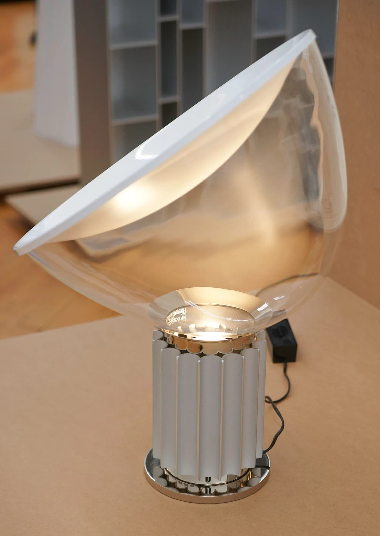 A Taccia lamp with a large clear glass bowl tilted on a ribbed metal base, illuminating its white reflector above a pale floor.