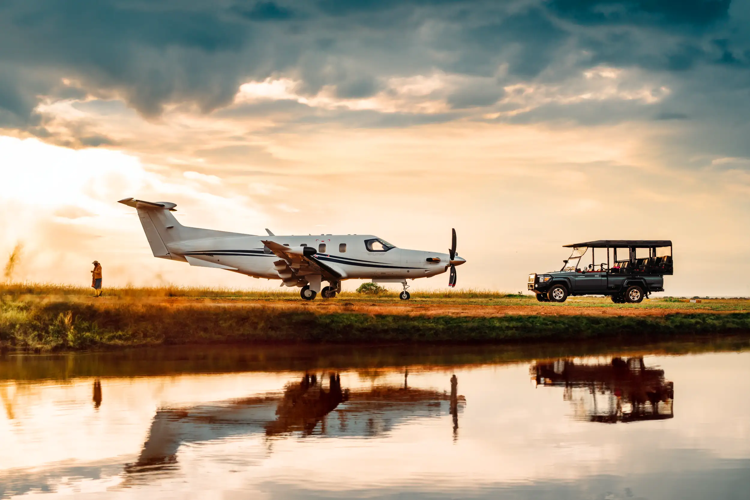 A white Pilatus PC-12 PRO on an unpaved strip beside a safari vehicle under a dramatic sky.