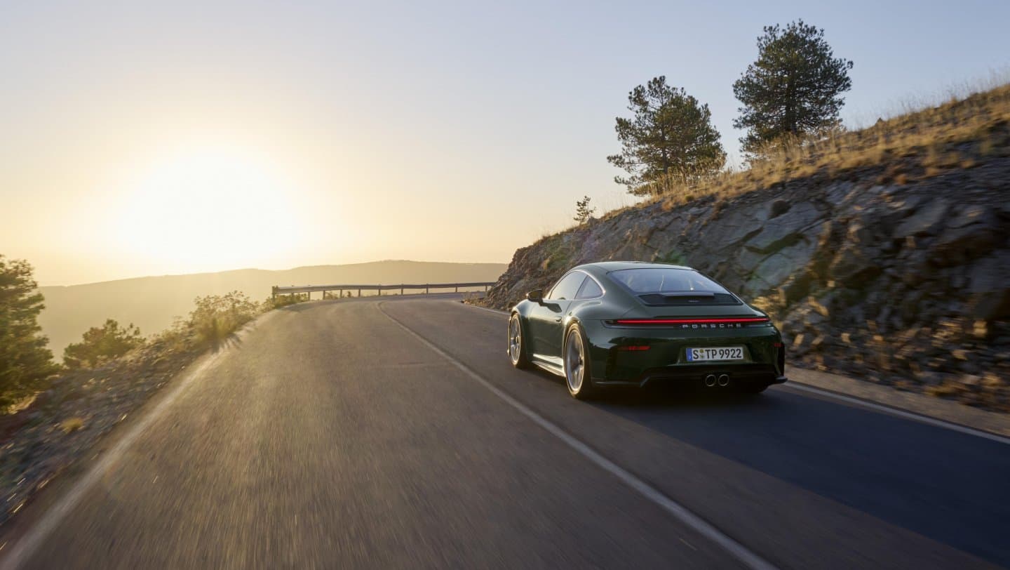A dark green 2025 Porsche 911 GT3 with Touring package driving on a mountain road in low evening light.
