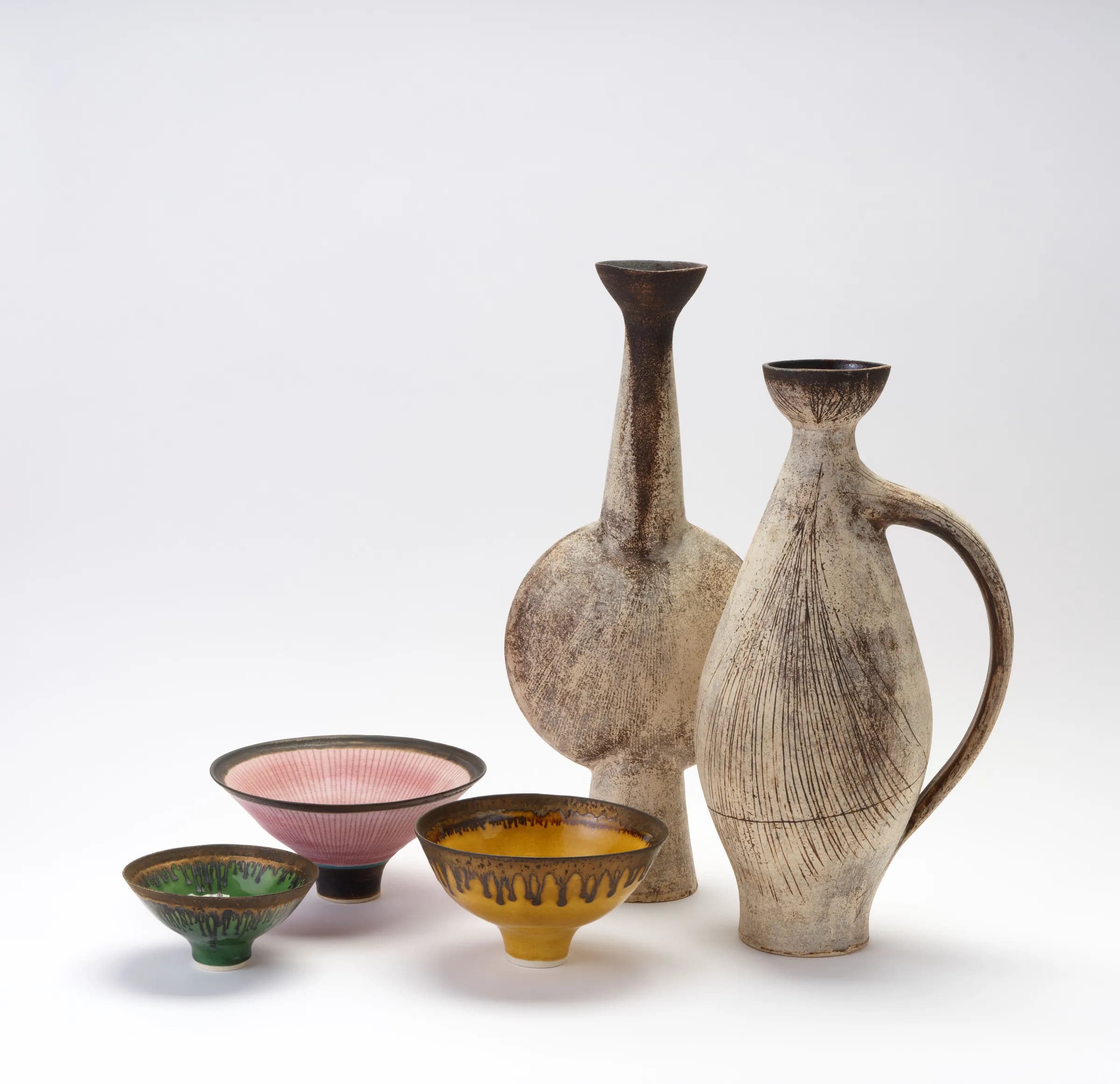 A group of bowls and tall ceramic vessels by Lucie Rie and Hans Coper against a pale background.