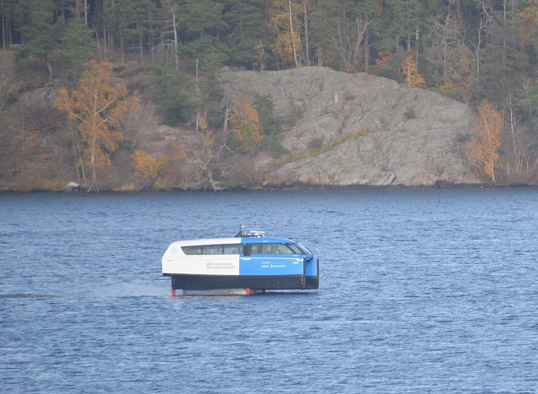 A blue-and-white Candela P-12 Shuttle hydrofoil ferry seen on the water during testing, with a rocky and autumn-coloured Stockholm shoreline behind it.