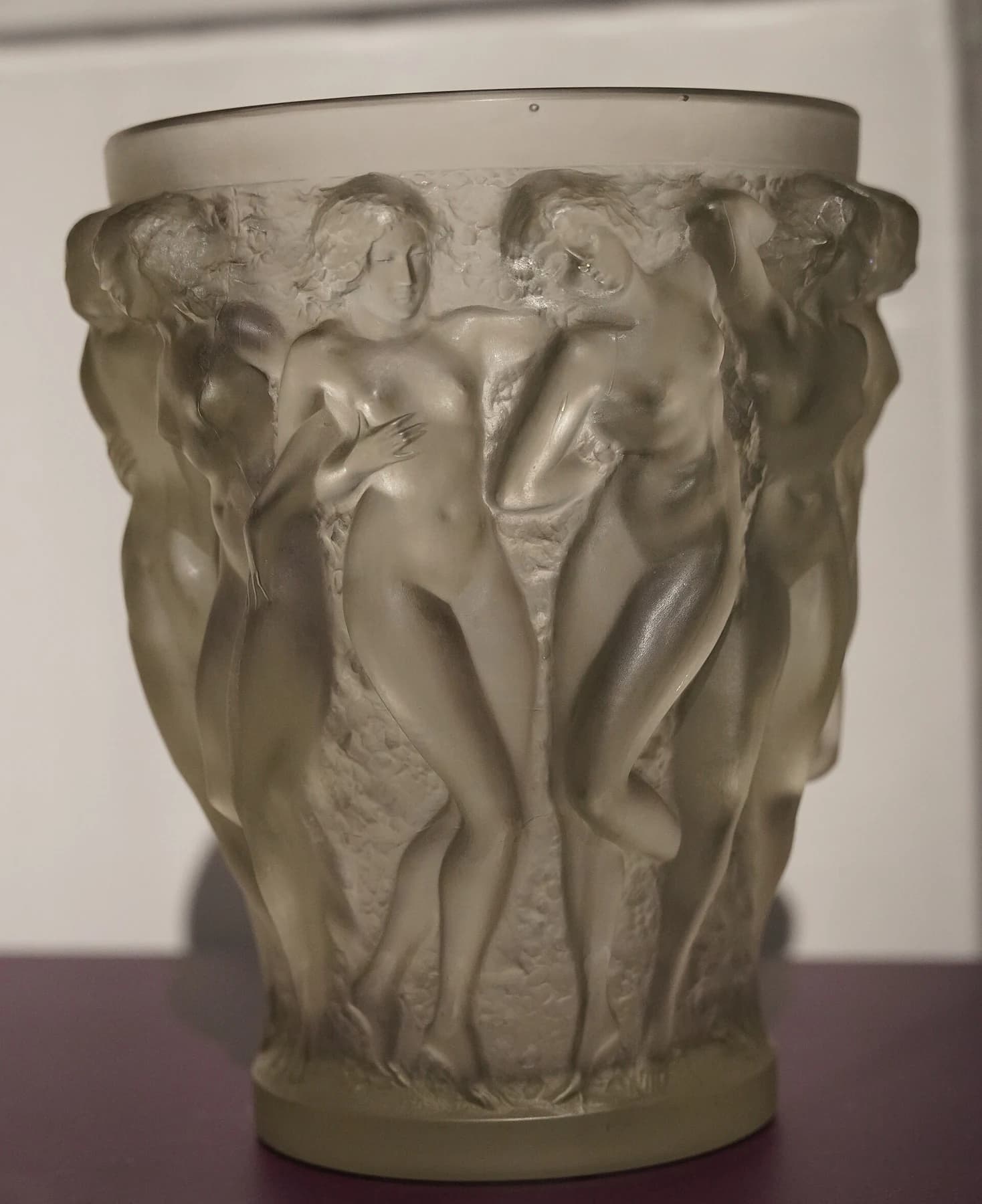A historical Lalique Bacchantes vase in a museum display, with a continuous relief of dancing figures and contrasting translucent and satin-textured glass.