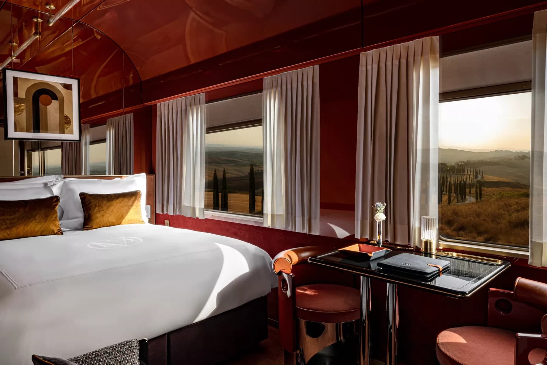 A La Dolce Vita Orient Express cabin with warm red wall panels, a bed, a small dark table and broad windows looking towards a Tuscan-style landscape.