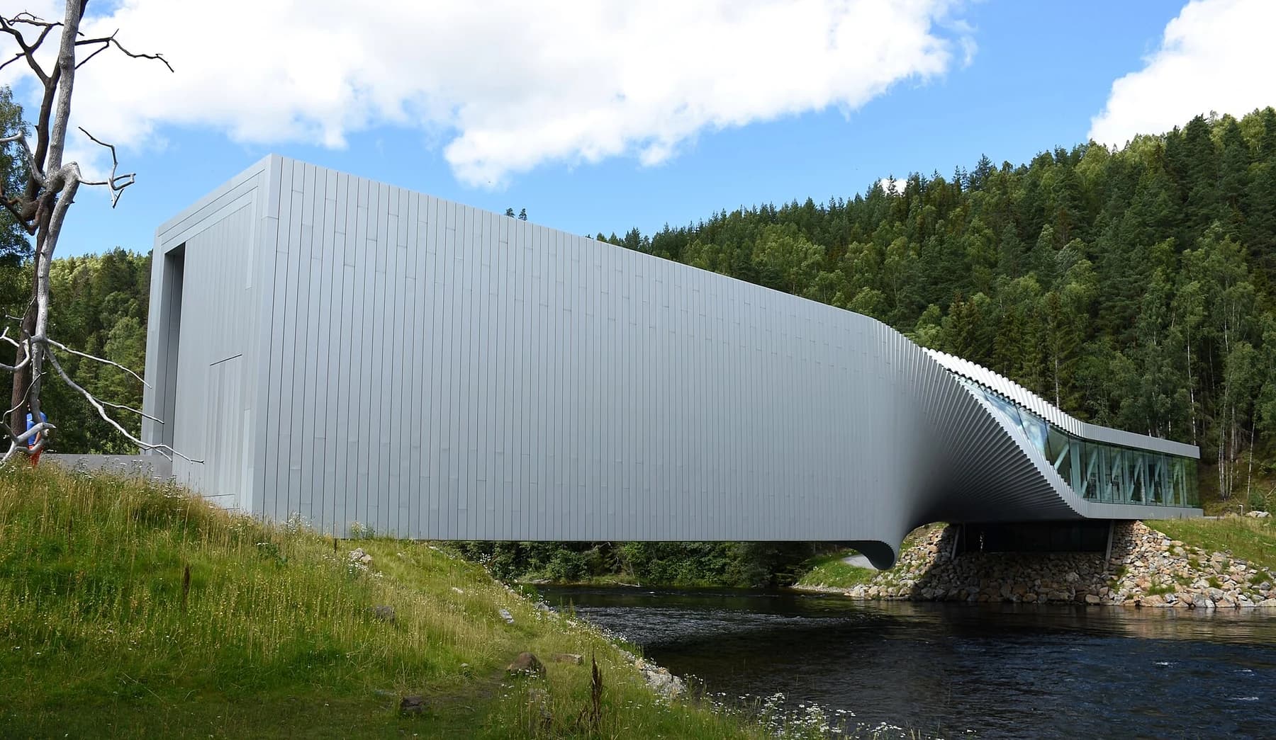 The pale twisting gallery bridge at Kistefos spans the Randselva between green riverbanks beneath a bright blue and clouded sky.