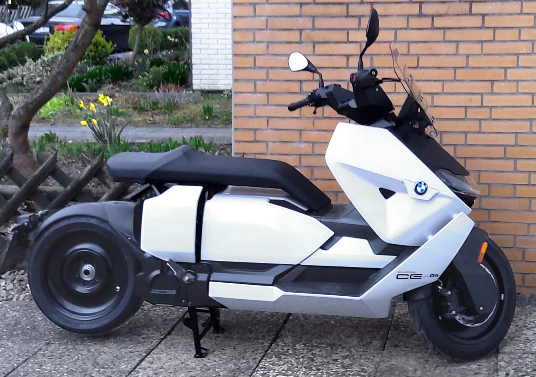 A white and black BMW CE 04 viewed from the side beside a brick wall, showing its long seat, low body, wheels and windscreen.