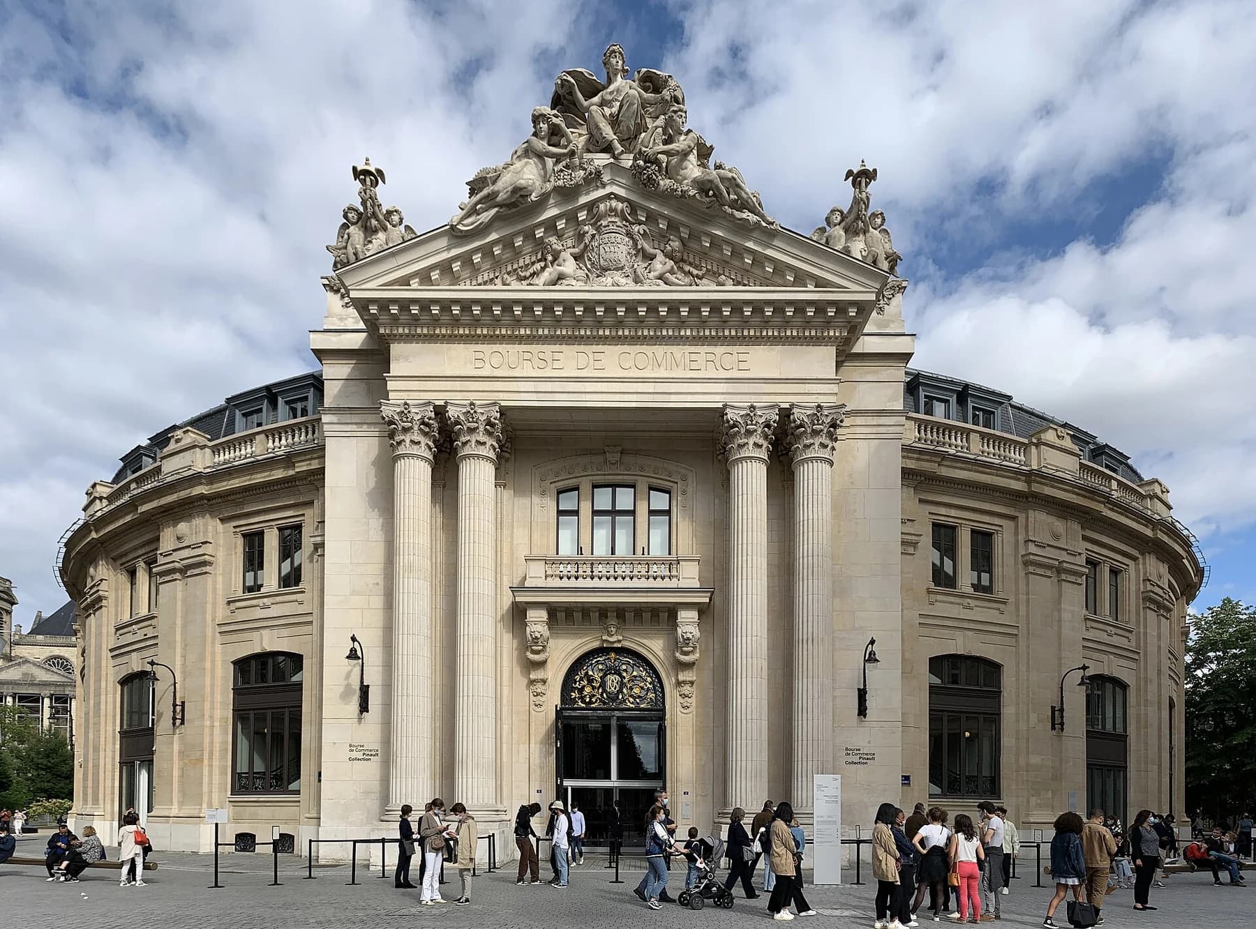 The curved historical exterior and sculpted entrance pediment of the Bourse de Commerce in Paris, with visitors in the square under a partly cloudy sky.