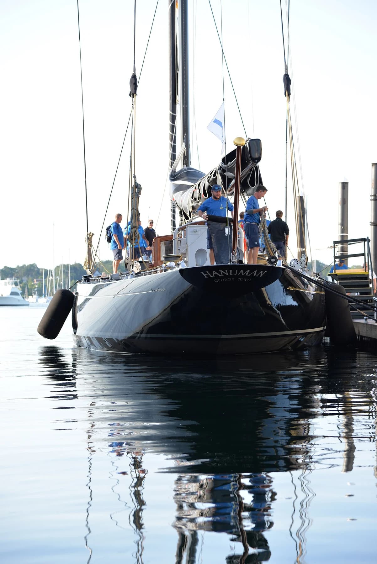 Hanuman seen from the stern at a dock with crew on deck, its name visible in pale lettering above a dark hull reflected in still water.