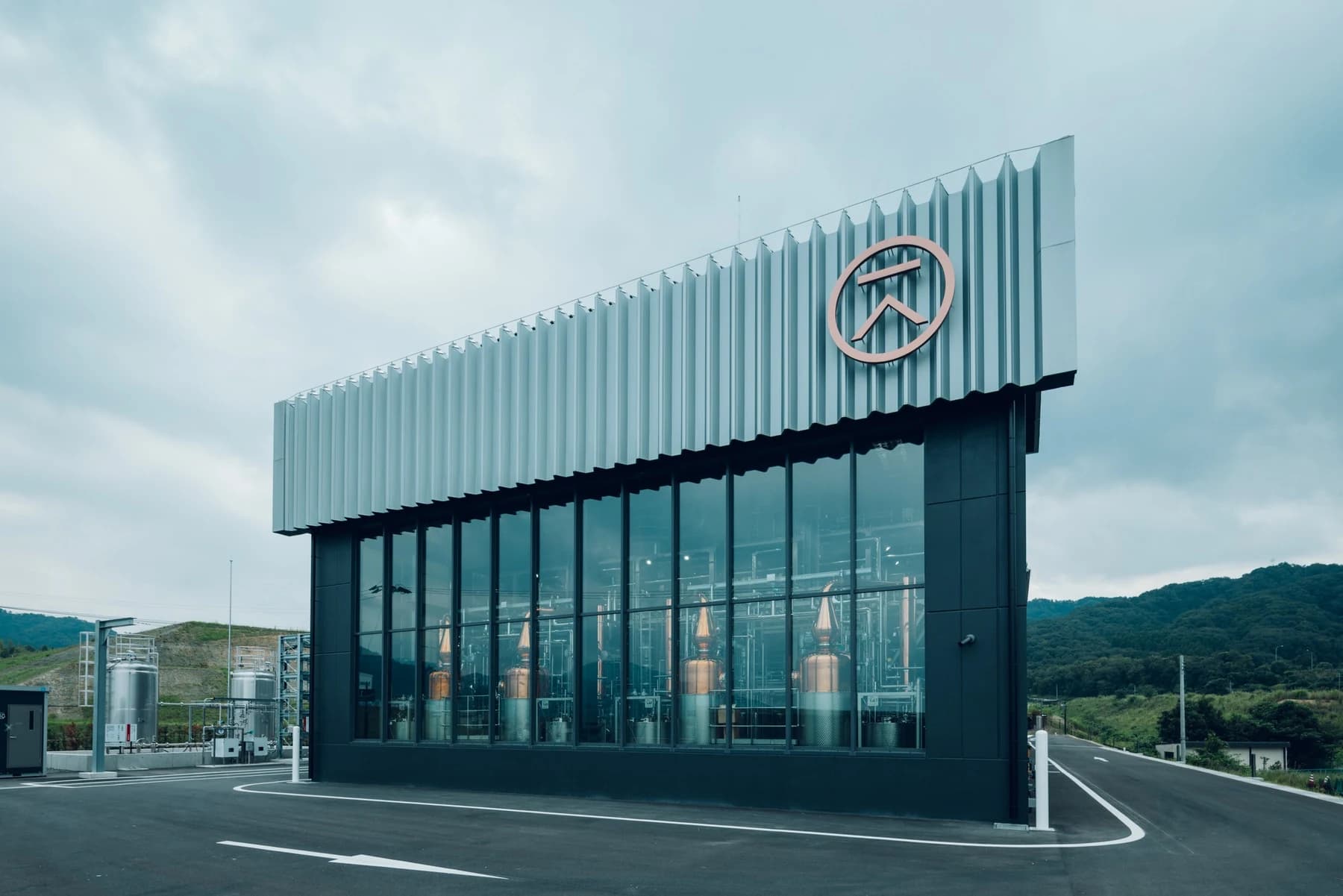 The new KI NO BI distillery at Kameoka with copper stills visible through tall front windows beneath a ridged metal façade and the brand emblem.