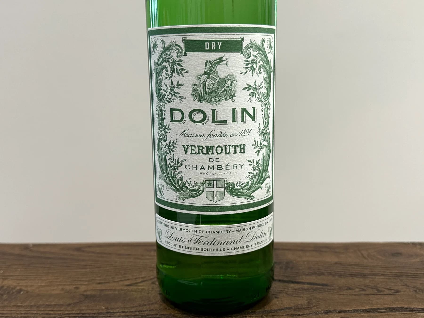 A green Dolin Dry vermouth bottle with its ornate cream-and-green Chambéry label standing on a wooden surface.