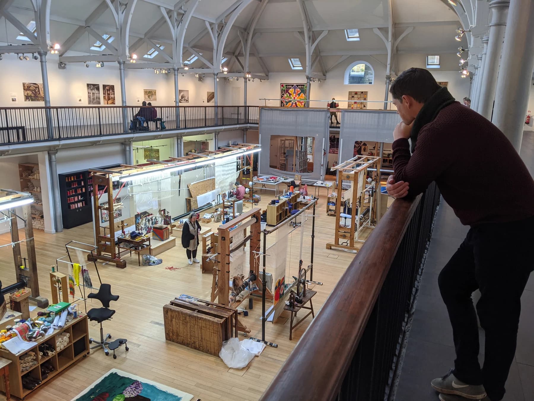 Dovecot’s large weaving room seen from a balcony, with looms and work areas below a high white roof and visitors looking down from the railing.