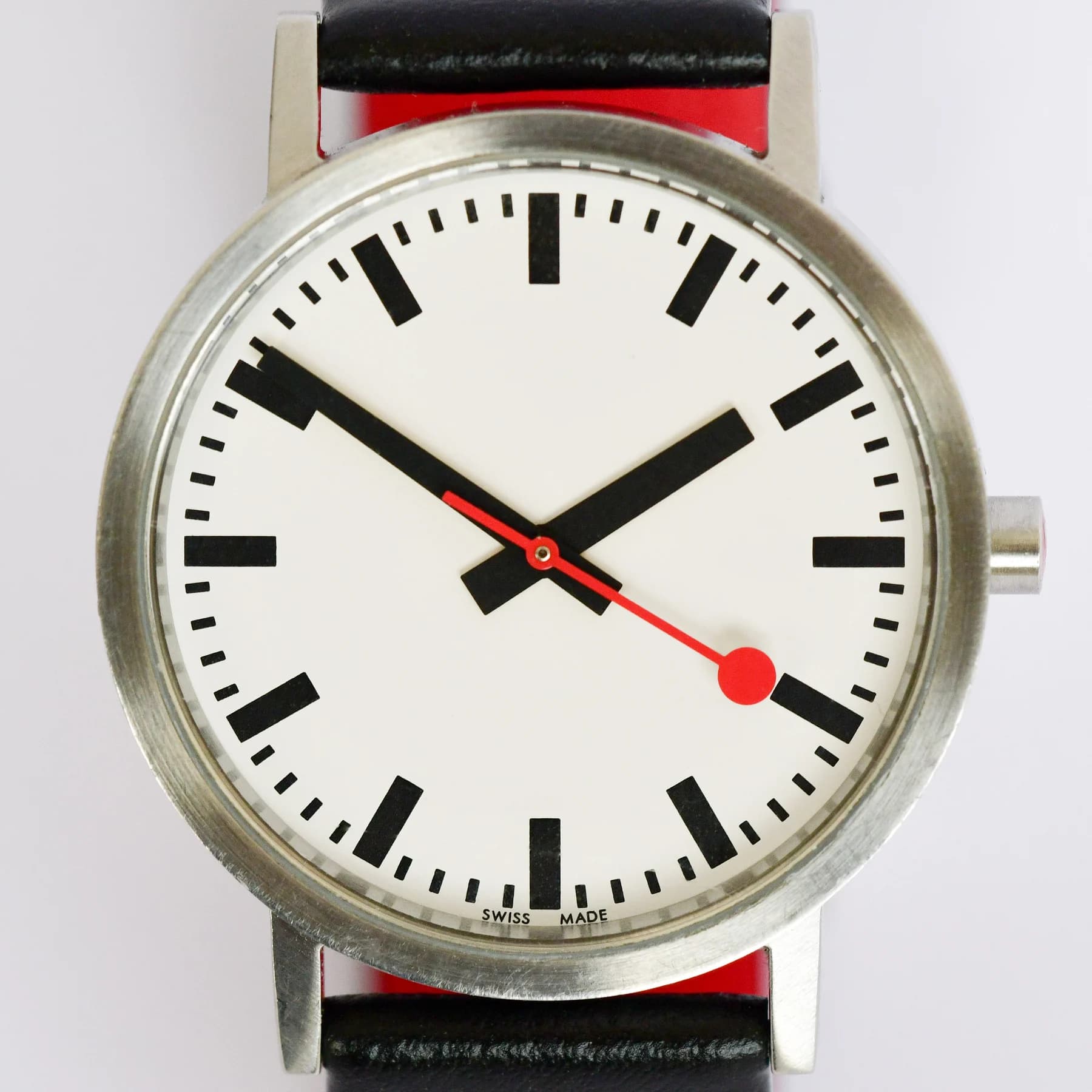 A Mondaine watch with a white dial, bold black hour marks and hands, a red round-tipped seconds hand and a red-and-black strap.