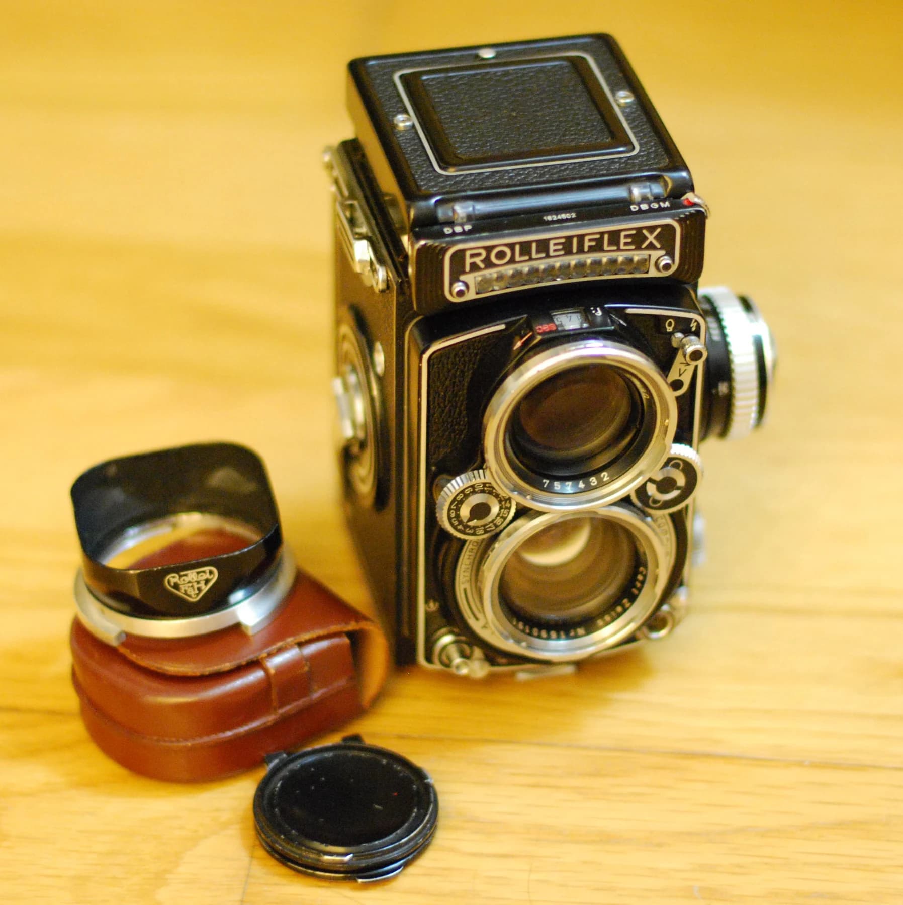 A Rolleiflex twin-lens camera with two vertically arranged lenses stands on a warm wooden surface beside its case and lens accessories.