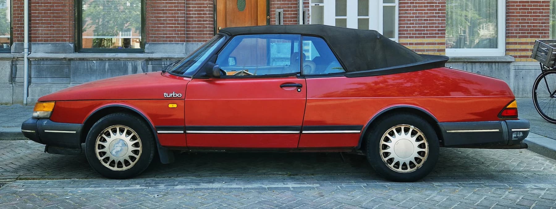 A red classic Saab 900 Turbo convertible with its black fabric roof raised, photographed in side profile beside a brick building.