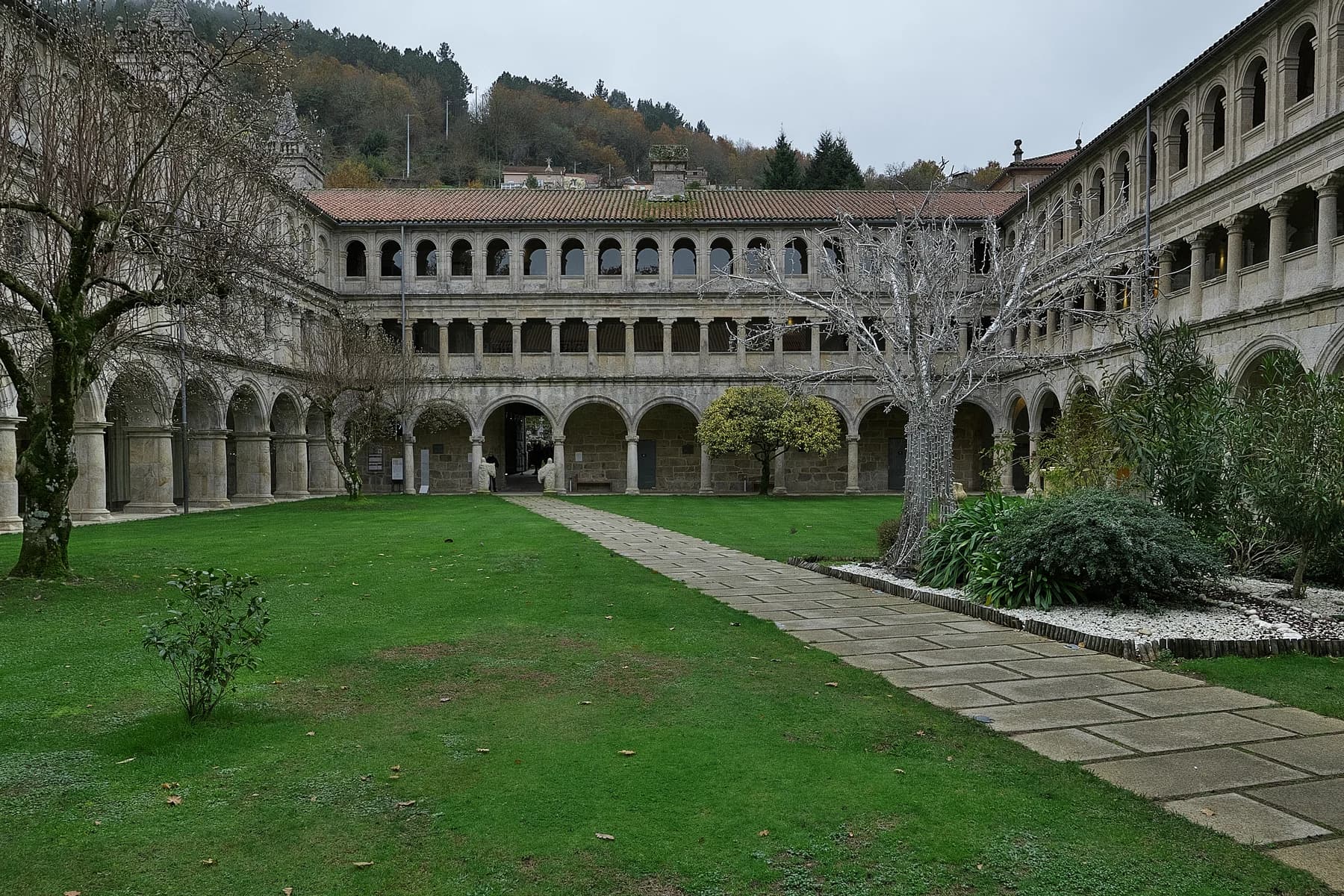 Arcaded stone galleries surround a green courtyard with trees and paths at the monastery of Santo Estevo de Ribas de Sil.