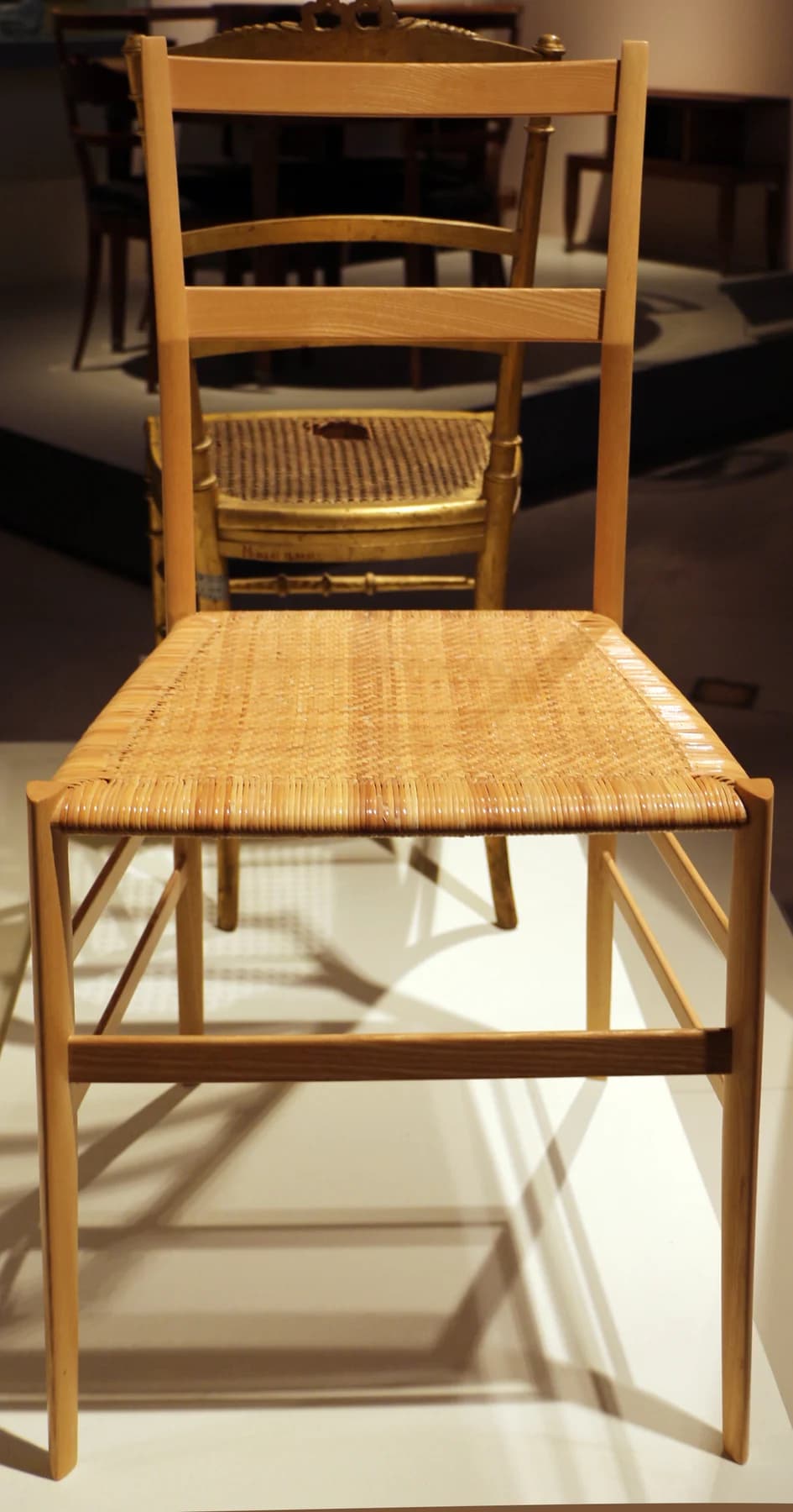 A slender wooden Superleggera chair with a woven seat stands front-on in a museum display, with other chairs visible behind it.