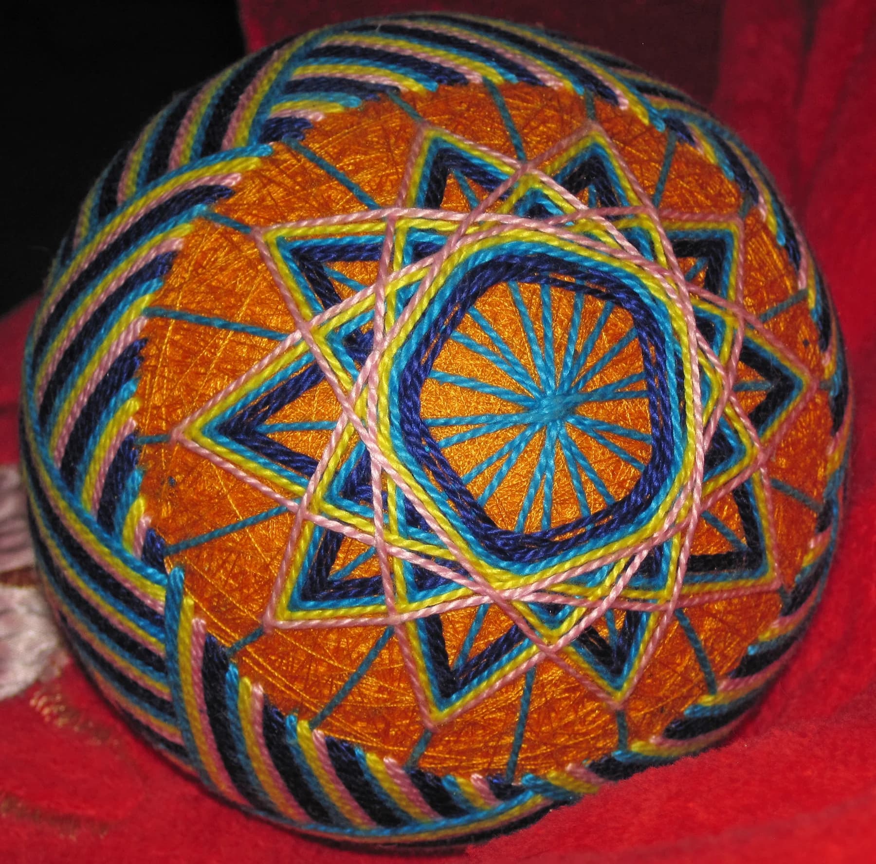 An orange temari ball covered with interlocking blue, green and pale geometric stitching on a red surface.