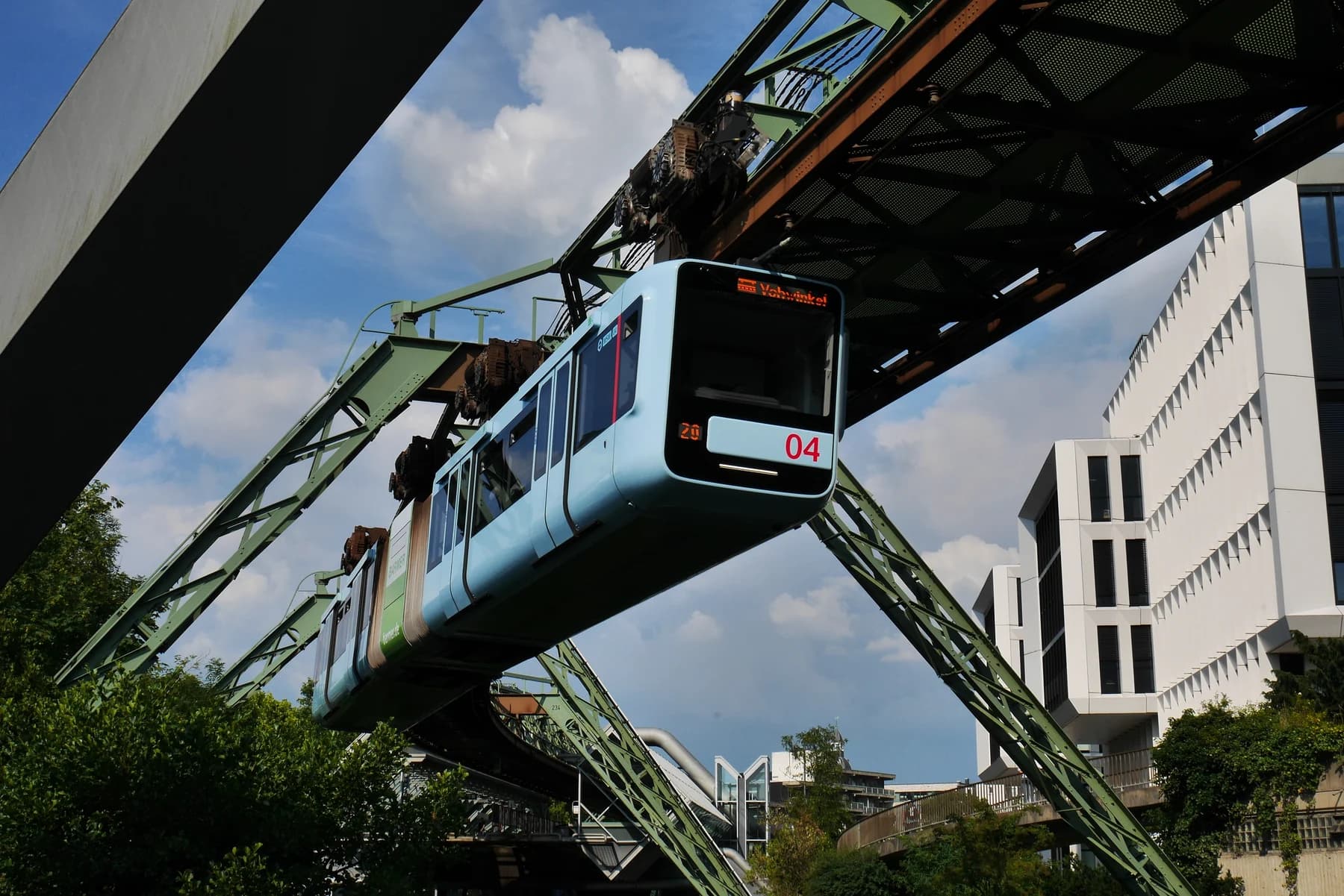A blue articulated Wuppertal Schwebebahn train hangs beneath a steel track above the river, framed by the city’s bridges and buildings.