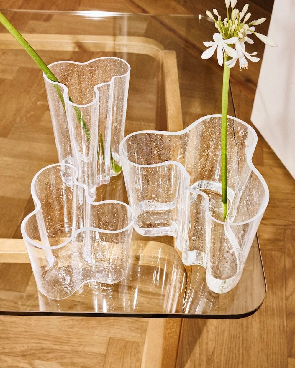 Three clear Aalto Bubble vases in different heights on a wooden surface, with flowers and branches revealing bubbles within the curved glass walls.