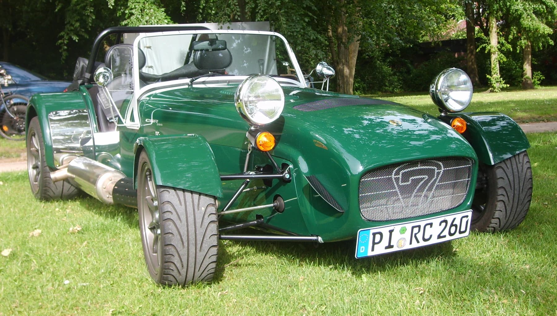 A green Caterham CSR 260 on grass at a vehicle event, seen from the front with its exposed wheels, suspension and compact open cockpit.