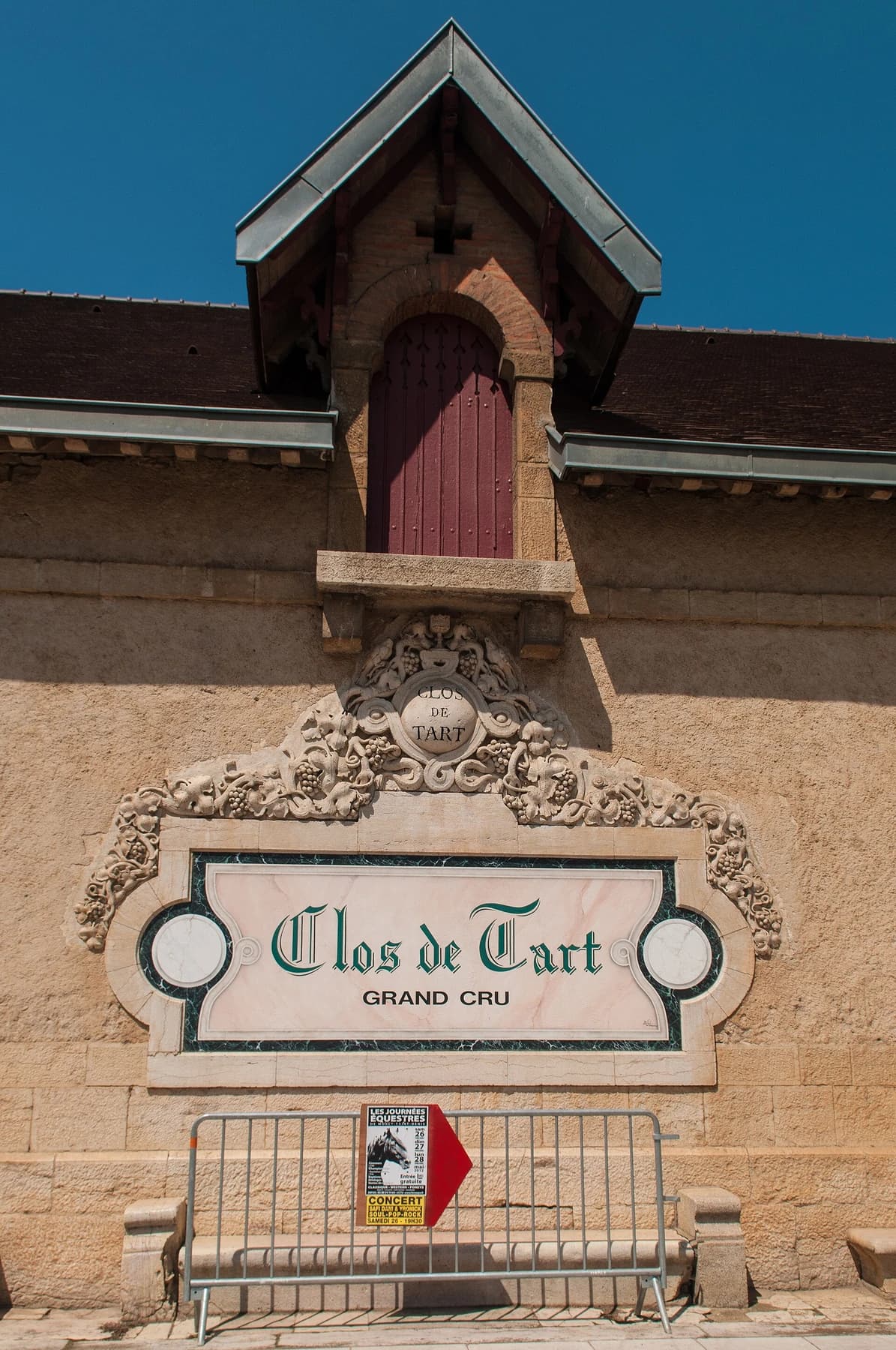 The stone facade and Grand Cru sign at Clos de Tart, with a red shuttered opening above the carved estate name against blue sky.