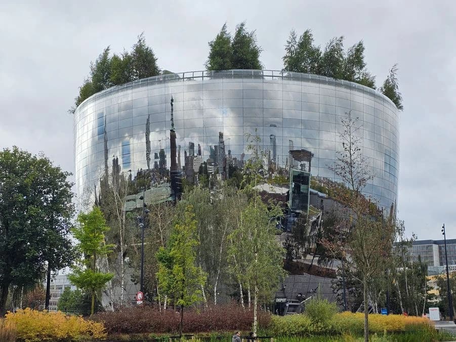The curved mirrored exterior of Depot Boijmans Van Beuningen reflecting Rotterdam, with trees on its roof and planting in the foreground.