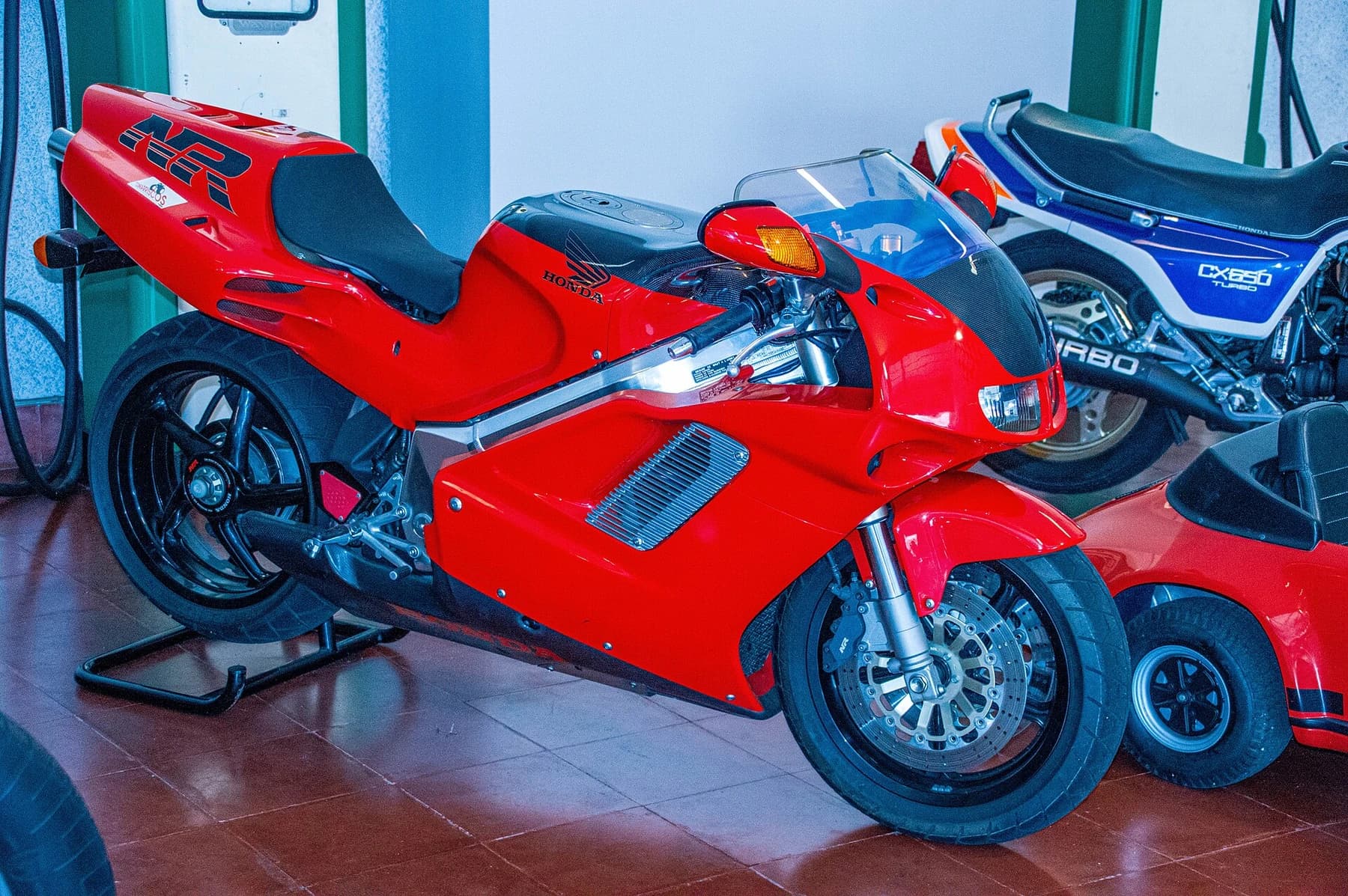 A red Honda NR750 RC40 displayed indoors in front of other motorcycles, showing its enclosed bodywork and distinctive side openings.
