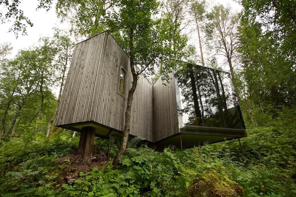 A timber-clad Landscape Room at Juvet, set on sloping woodland ground with a large glass wall reflecting trees beside the structure.