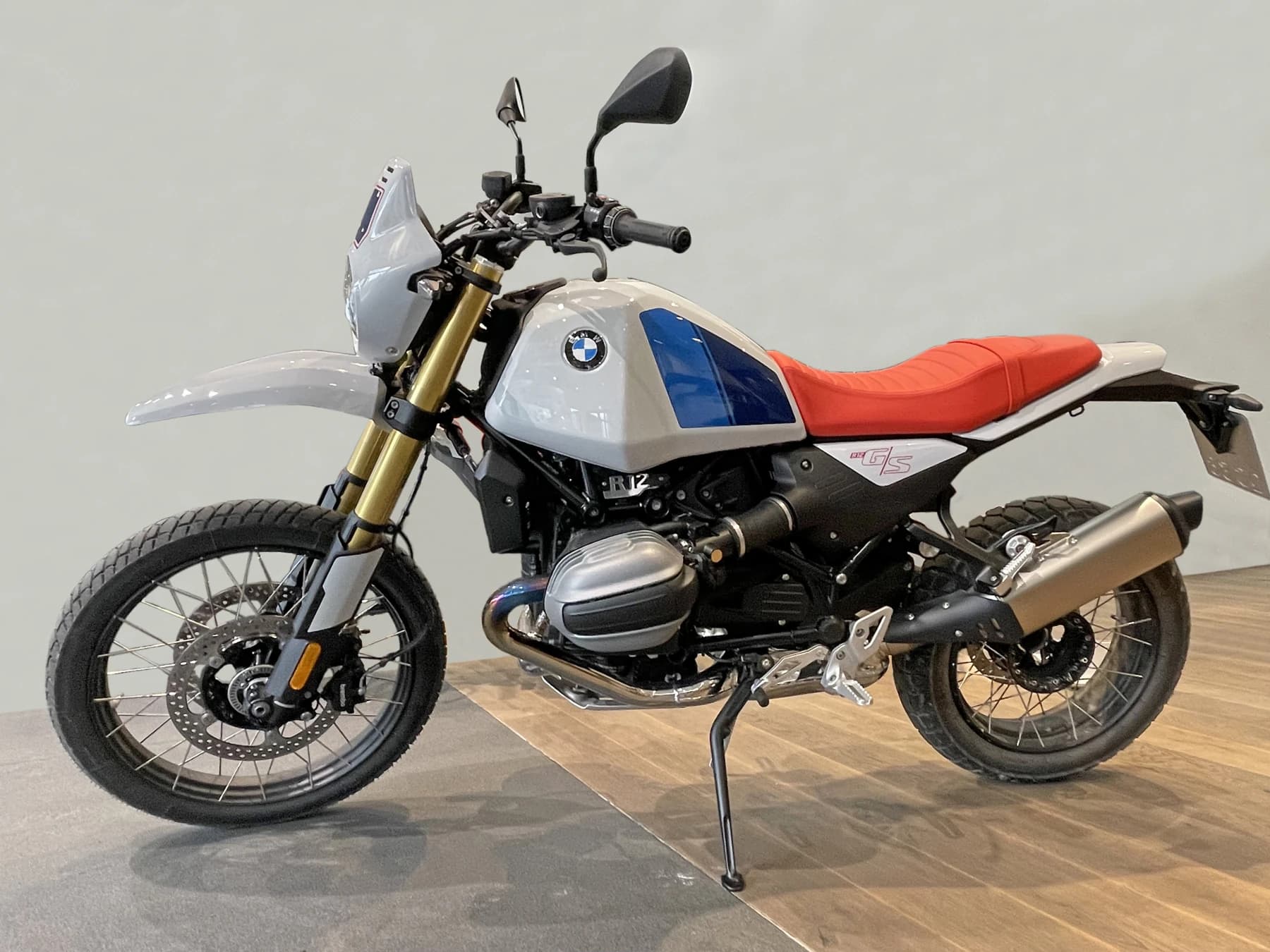 A 2025 BMW R 12 G/S motorcycle in white, blue and orange, showing its boxer engine, raised stance and spoked wheels against a light background.