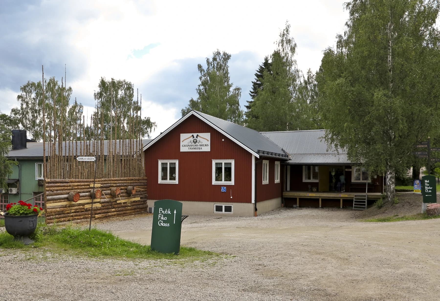The red timber exterior of Gränsfors Bruk’s axe forge in Sweden, with its sign, stacked timber, trees and gravel approach visible in daylight.