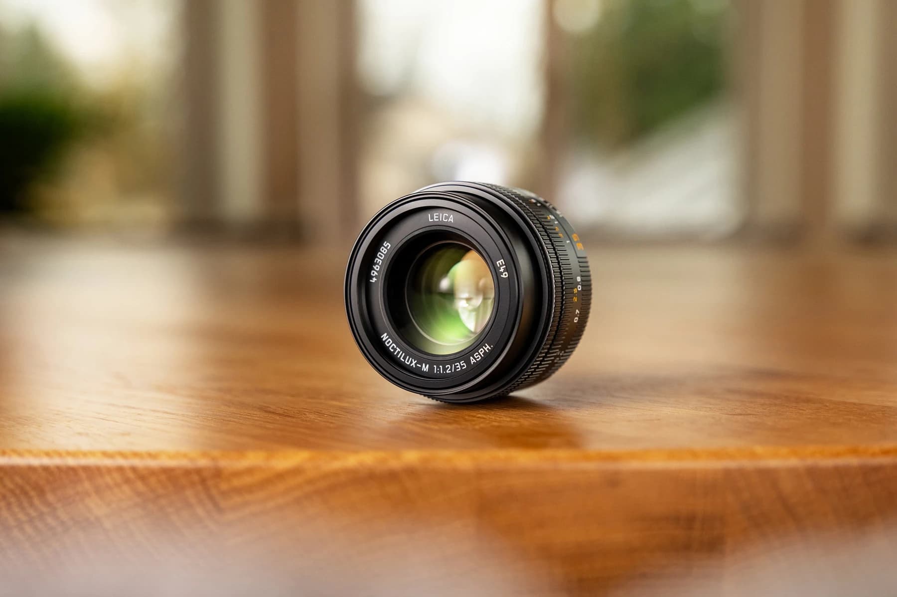 The black Leica Noctilux-M 35 f/1.2 ASPH. lens resting on a warm wooden surface, with the front glass and lens markings visible against a softly blurred background.