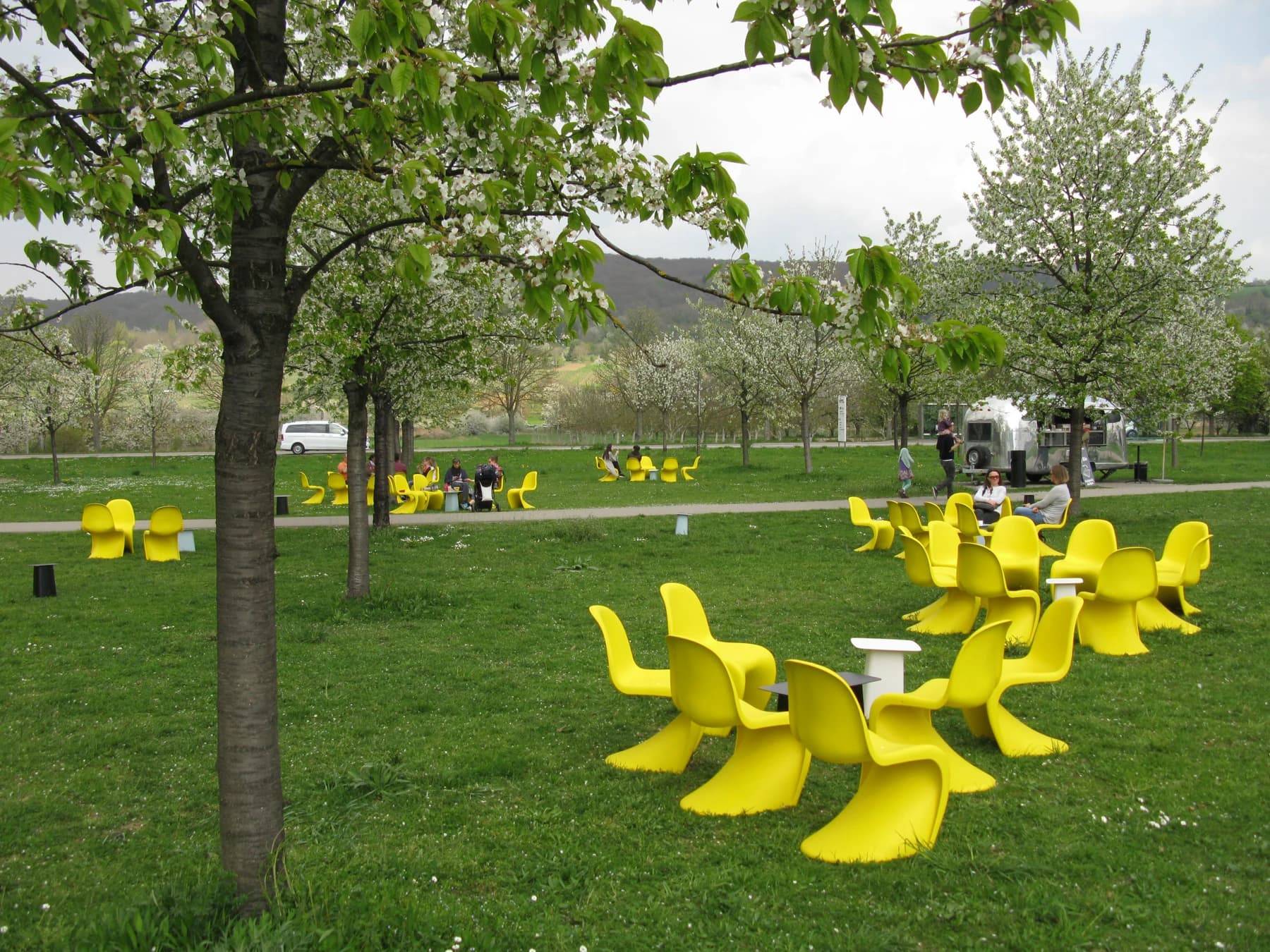 Yellow Panton Chairs arranged beneath trees outside the VitraHaus in Weil am Rhein, with tables and an Airstream kiosk behind the seating.