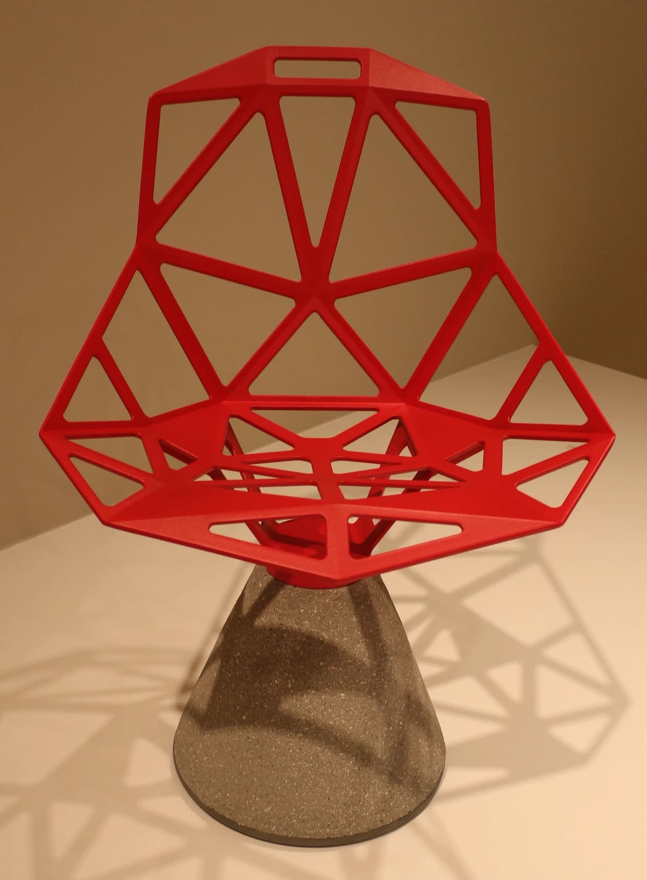A red Chair_One with an open triangular aluminium seat on a tapered concrete base, casting angular shadows on a pale display platform.