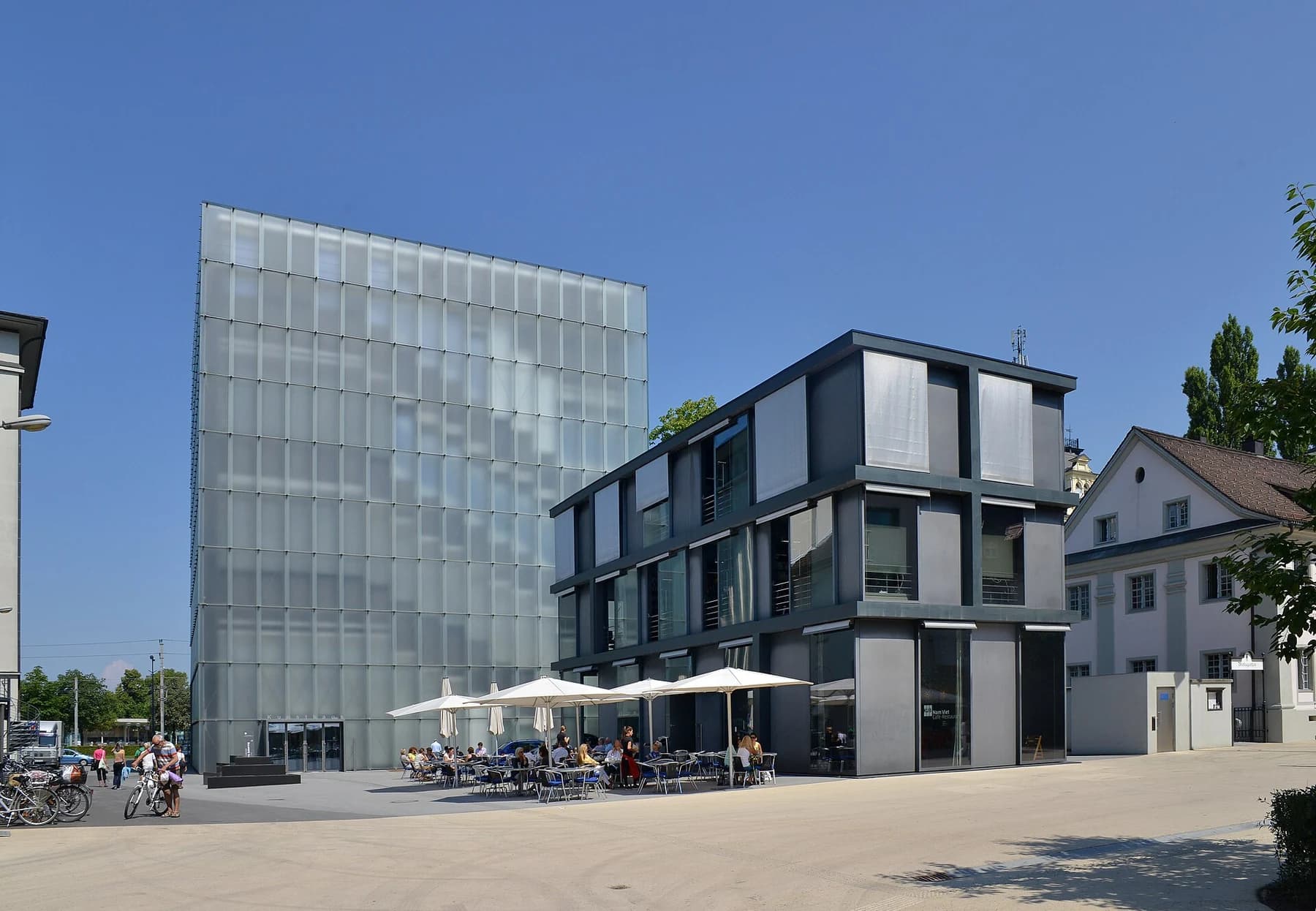 The translucent glass volume of Kunsthaus Bregenz behind its lower adjoining building, seen across a square under a clear blue sky.