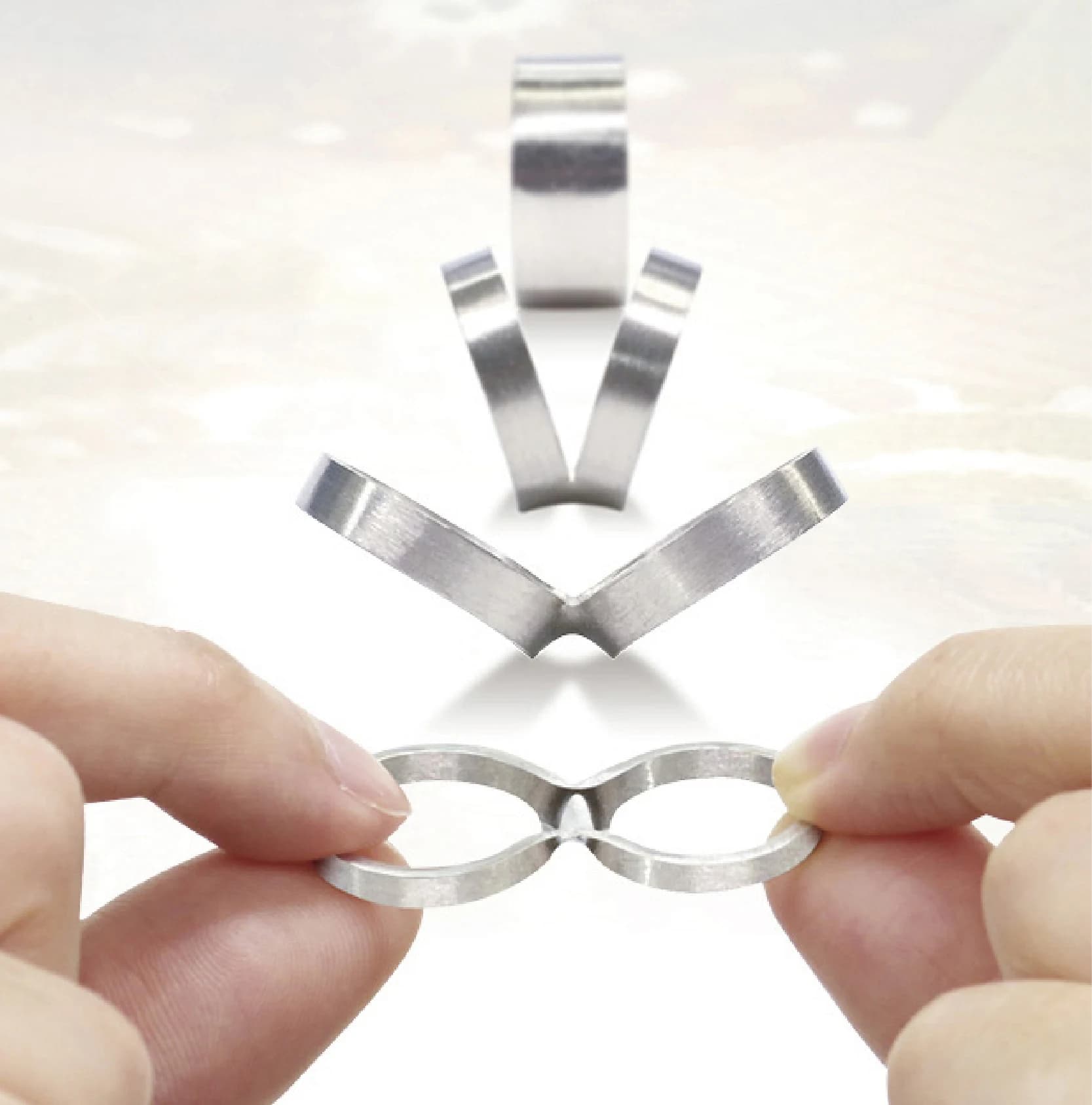 Hands holding two connected rings, with earlier stages of the metal band opening into a pair arranged behind them.