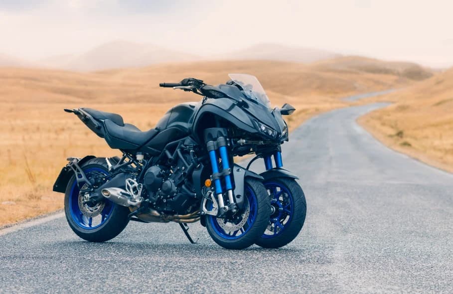 A dark Yamaha Niken on a winding road through golden grass, viewed from the front quarter with both blue-rimmed front wheels clearly visible.