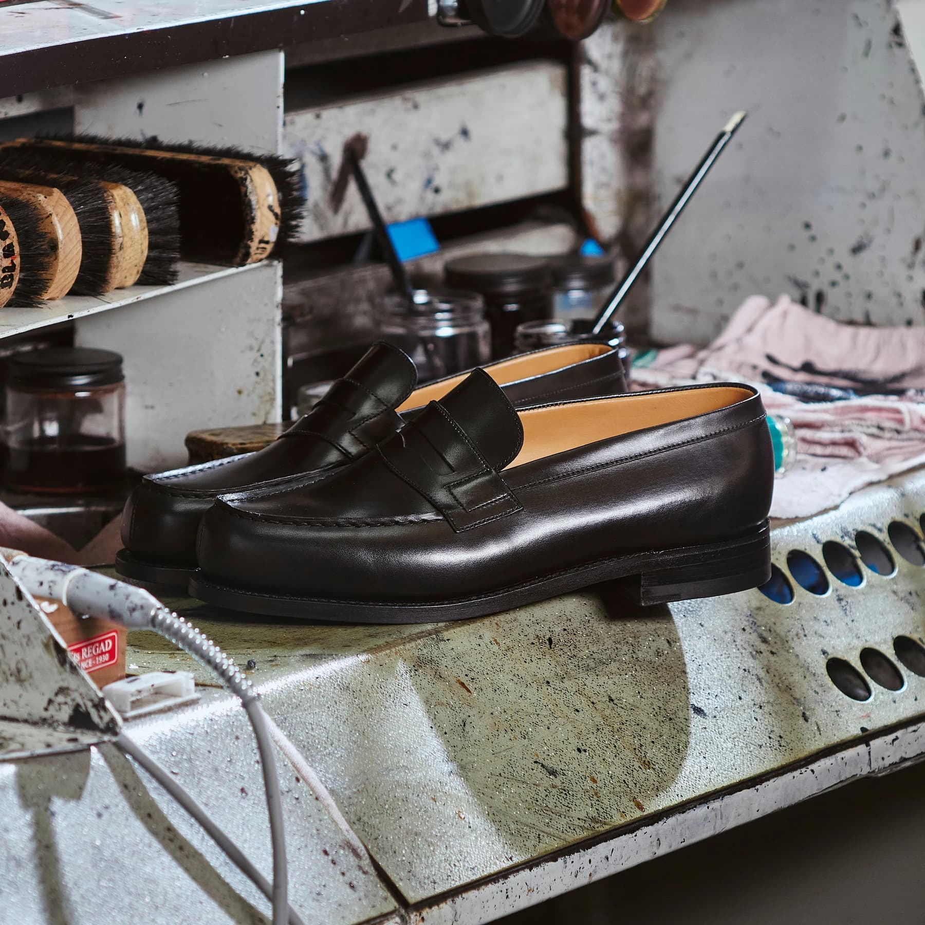 A polished black J.M. Weston 180 loafer resting on a workshop bench among tools, brushes and shoe-care materials.