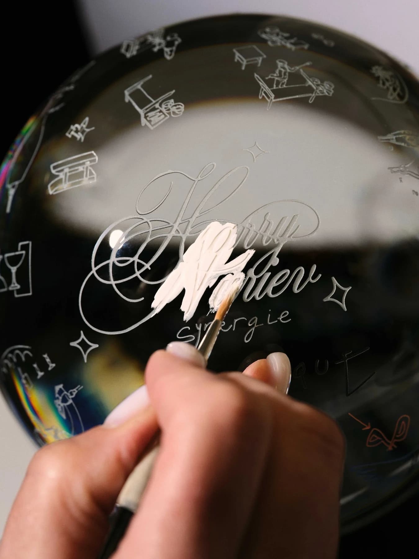 A close workshop photograph of a hand applying pale painted marks to a curved Baccarat Sirius crystal form bearing Harry Nuriev’s graphic words and small drawings.