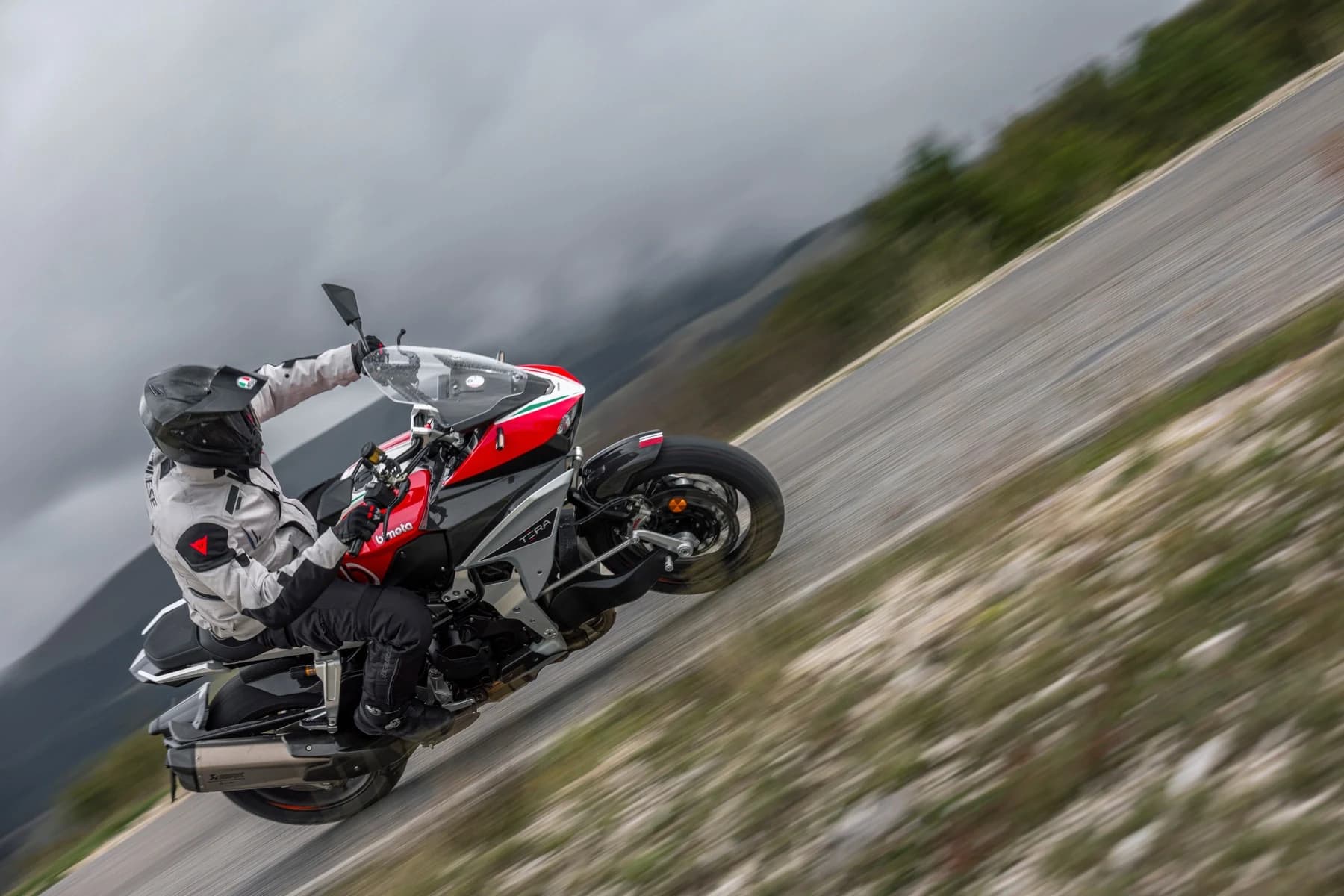 A helmeted rider leaning a red, white and carbon-finished Bimota Tesi H2 TERA along a road with a mountain landscape behind, in the supplied riding photograph.
