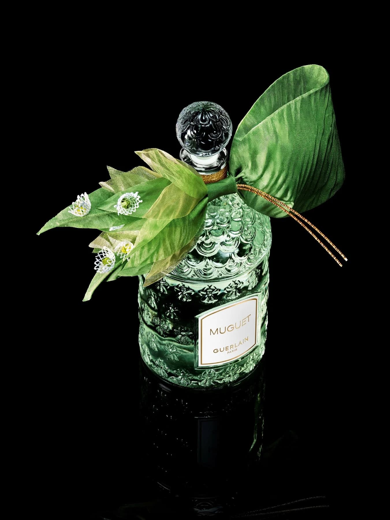 Guerlain Muguet 2026 bee bottle against black, decorated with a bright green pleated silk ribbon, embroidered white lily-of-the-valley bells and a clear stopper.