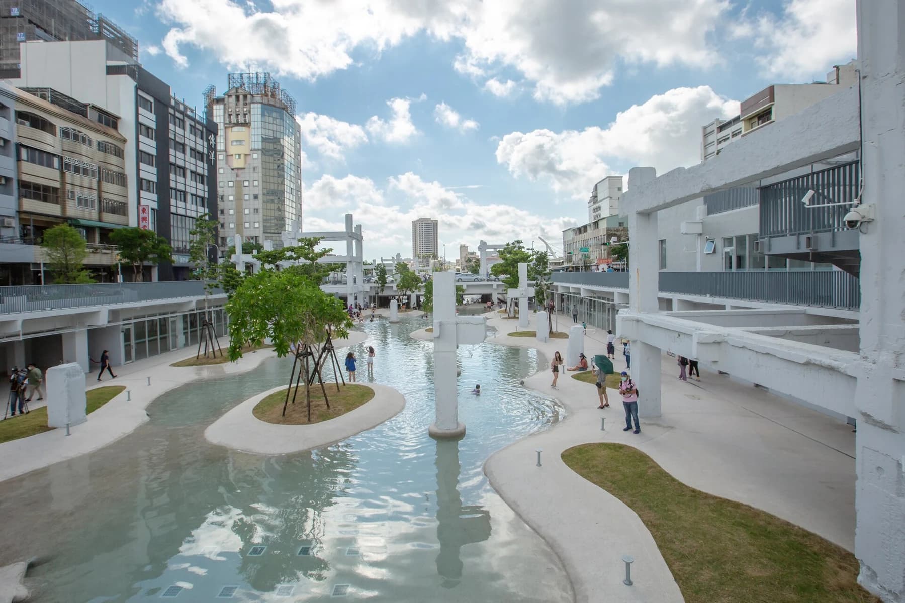 The sunken water landscape at Tainan Spring with curved planted islands, retained concrete columns, arcades and visitors, surrounded by urban buildings.