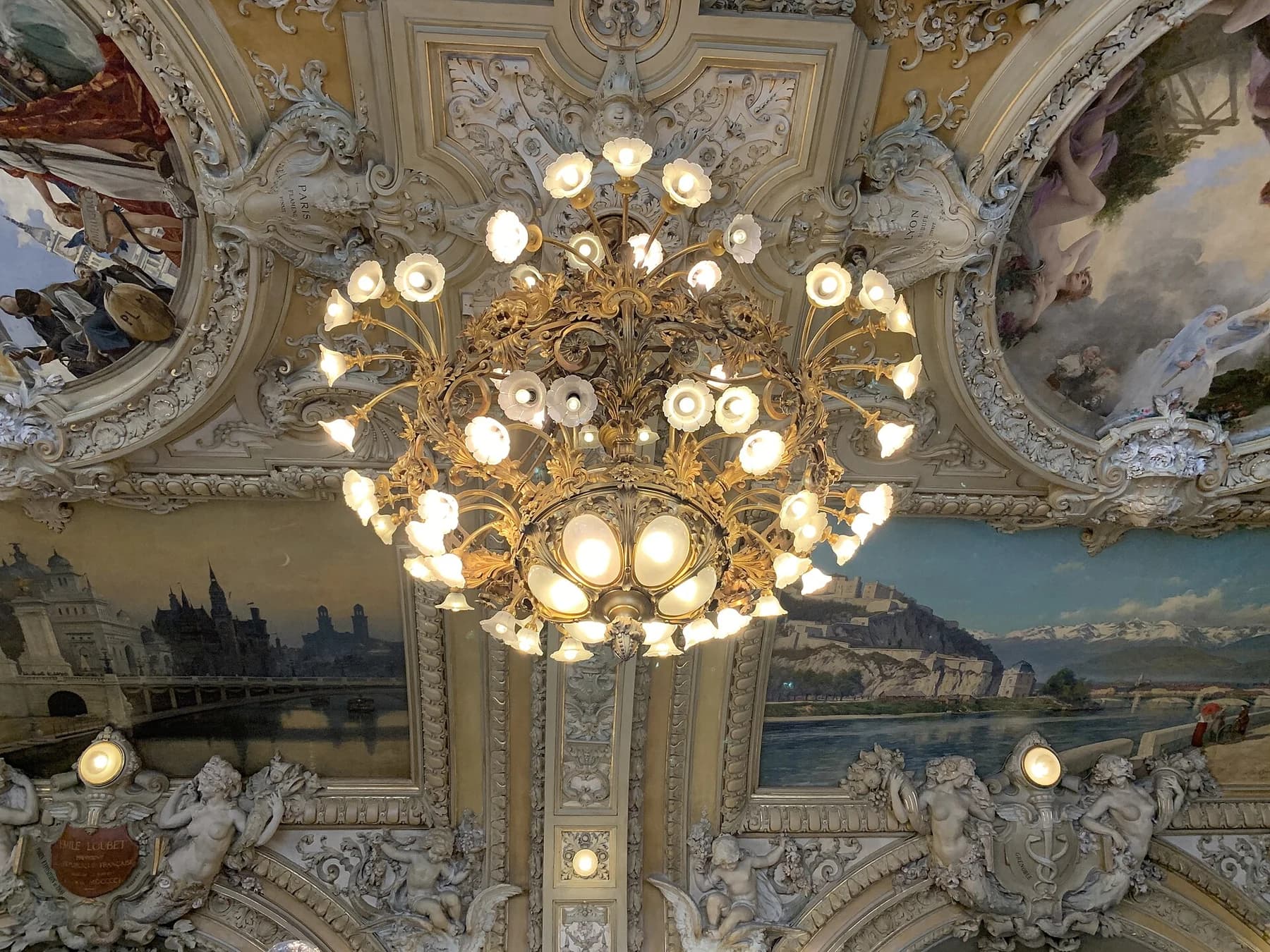 An ornate lit chandelier beneath the painted ceiling at Le Train Bleu in Gare de Lyon, Paris, with gilded architectural details and a painted landscape behind it.