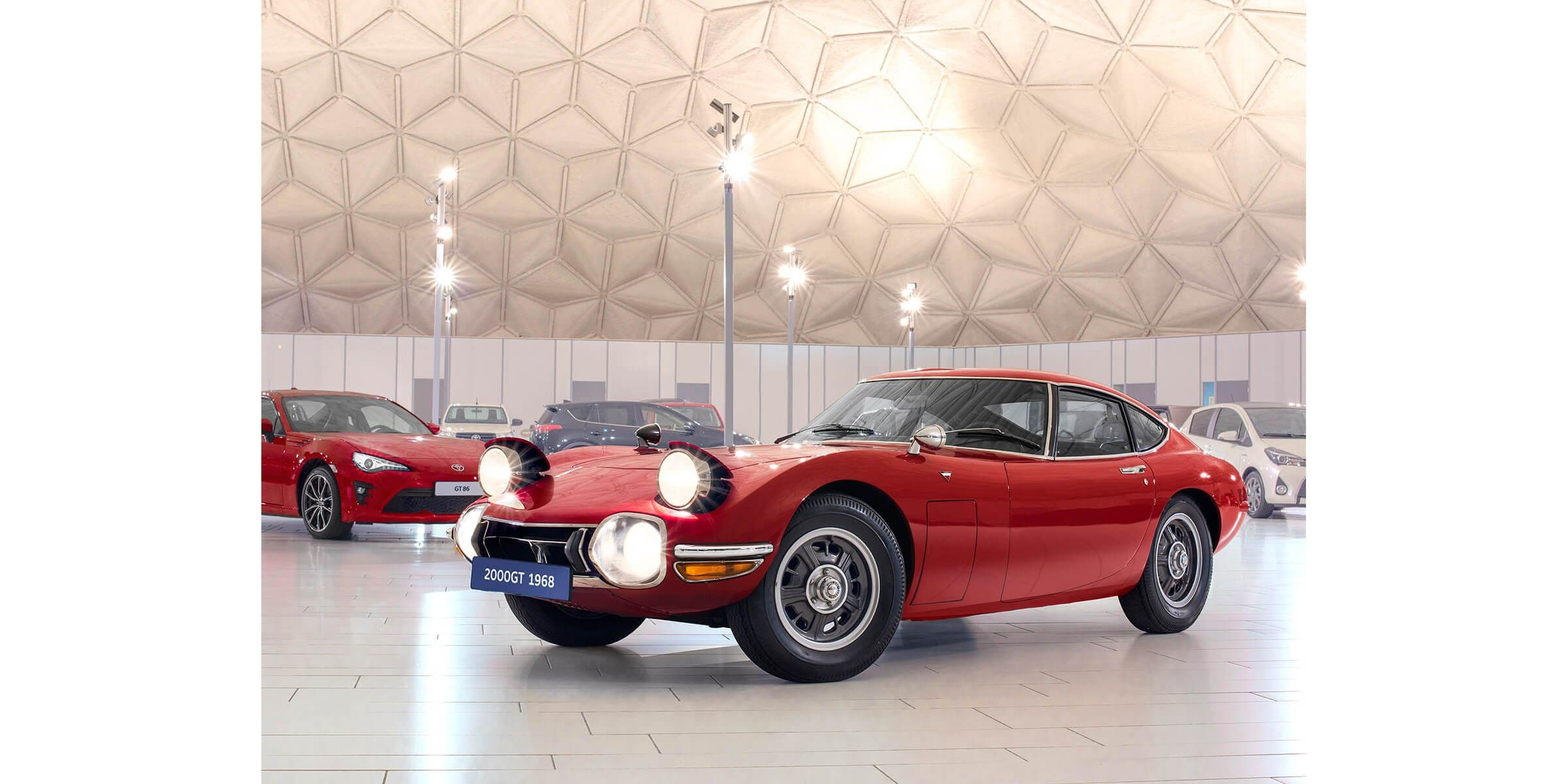 A red 1968 Toyota 2000GT shown from the front three-quarter angle with its headlamps raised inside a Toyota heritage display.