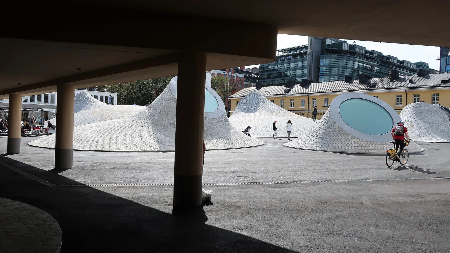 The pale domed roof forms of Amos Rex emerging into a Helsinki square, seen from the shade of a colonnade.