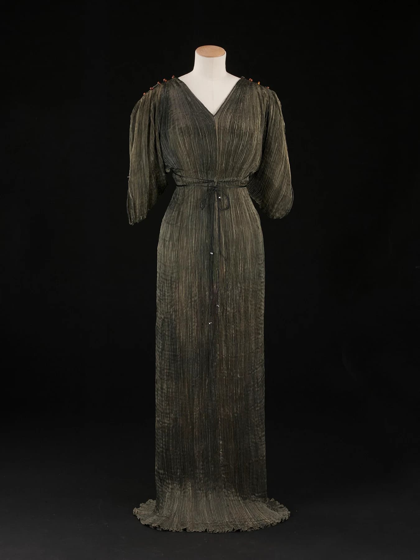 A long bronze-green Fortuny Delphos dress on a mannequin against black, with fine pleats, a V-shaped neckline and loose sleeves.