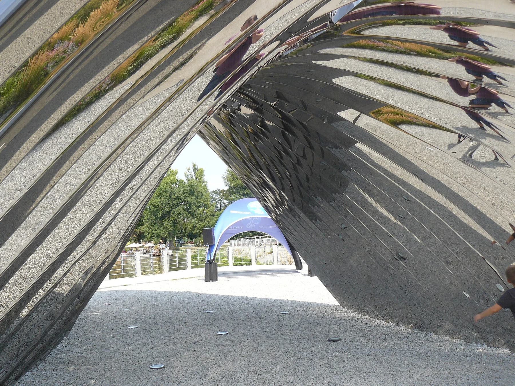 A close view through NAWA’s polished steel arches, reflecting the ground and people while framing trees on Wrocław’s Daliowa Island.